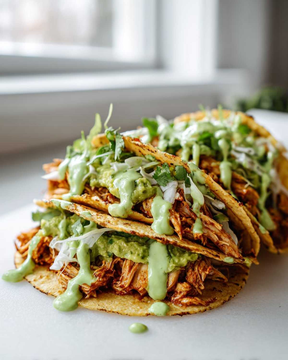 Three stacked, delicious Avocado Lime Crema Chicken Tacos filled with shredded chicken and topped with green sauce.