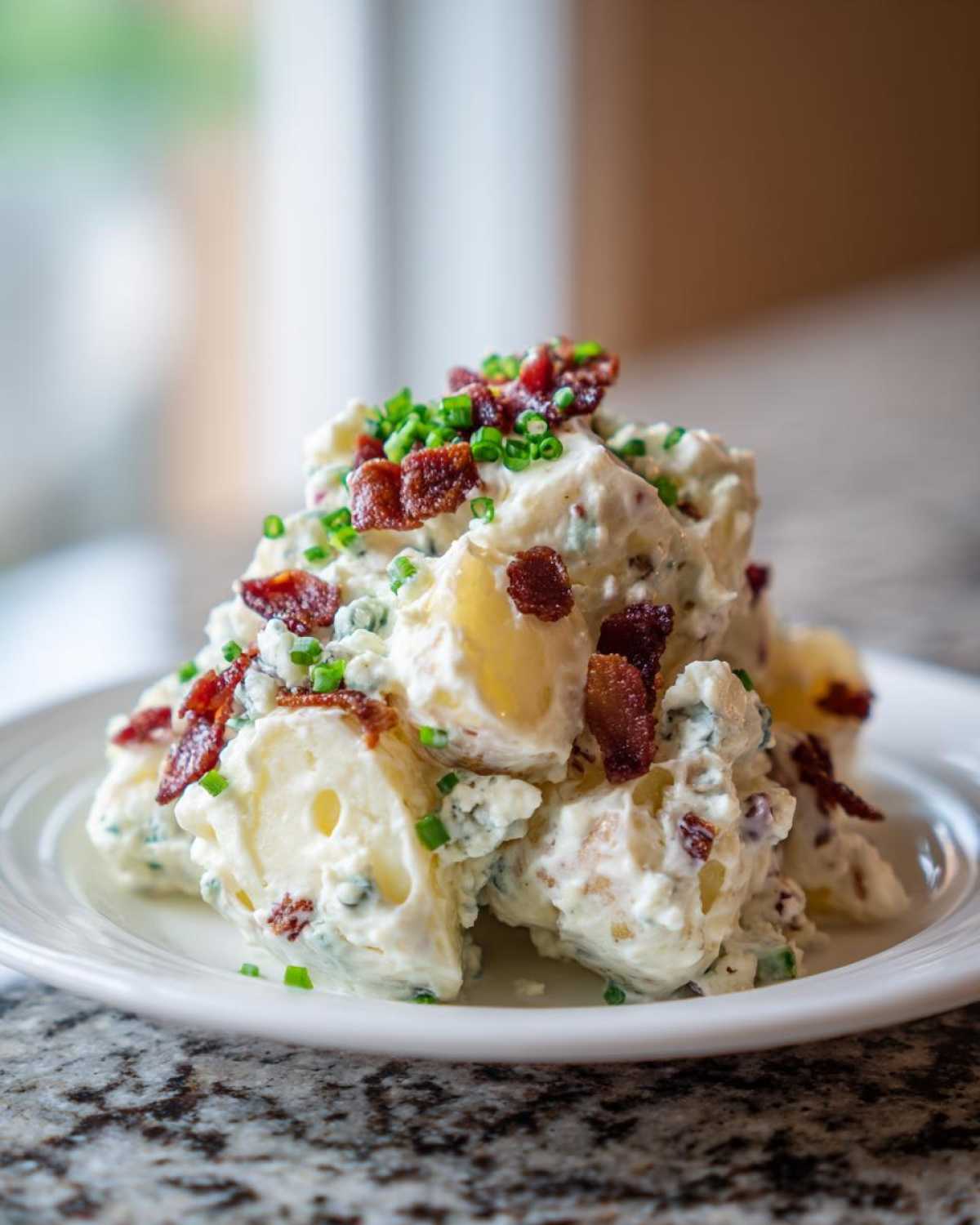 A mound of creamy Bacon & Blue Cheese Potato Salad topped with crispy bacon bits and fresh chives on a white plate.
