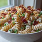 A close-up of creamy Bacon Ranch Pasta Memorial Day Salad featuring rotini pasta, green peas, bacon crumbles, and shredded cheese in a white bowl.