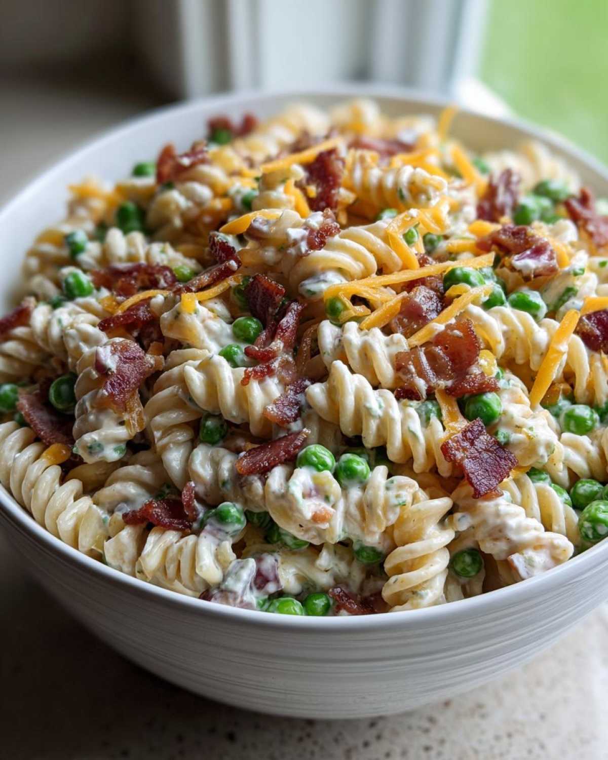 A close-up of creamy Bacon Ranch Pasta Salad featuring rotini pasta, green peas, crumbled bacon, and shredded cheddar cheese.