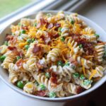 A close-up bowl of creamy Bacon Ranch Pasta Salad mixed with bright green peas, topped with shredded cheddar cheese and crispy bacon bits.