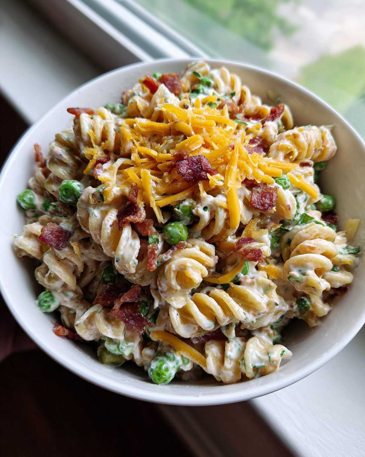 A close-up overhead shot of creamy Bacon Ranch Pasta Salad topped with shredded cheddar cheese and bacon bits in a white bowl.