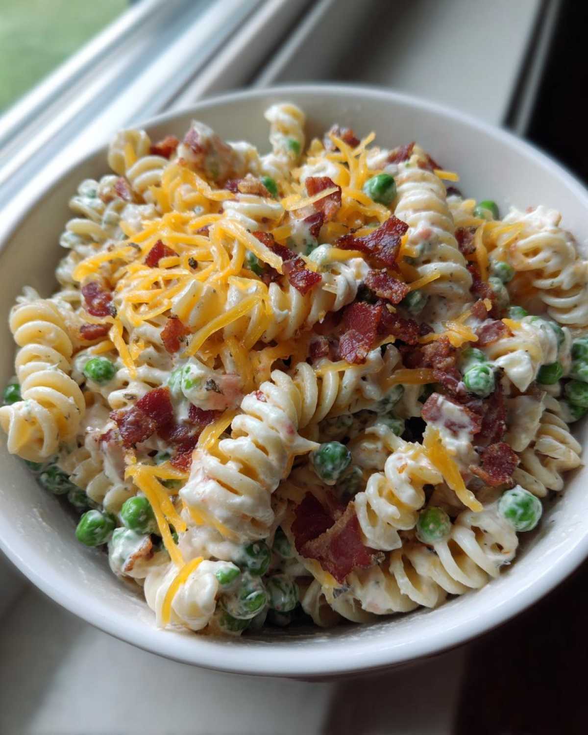 A close-up of a white bowl filled with creamy Bacon Ranch Pasta Salad mixed with green peas, topped with shredded cheddar cheese and crispy bacon bits.