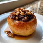 A close-up of a single, glistening Baked Apples with Cinnamon & Walnuts dessert on a white plate with syrup.