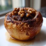 A close-up of a single, glistening Baked Apple with Cinnamon & Walnuts topping resting in syrup on a white plate.