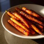A close-up of shiny, caramelized Balsamic Roasted Carrots piled on a white plate, catching the sunlight.