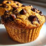 A close-up, sunlit shot of a freshly baked banana chocolate chip muffin topped with melted chocolate chips on a white plate.