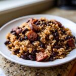 A close-up of a white plate piled high with seasoned beans and rice mixed with slices of smoked sausage.