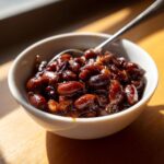 A small white bowl filled with rich, dark, saucy Baked Beans: The Best Baked Beans Ever, with a spoon resting inside.