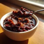 A spoonful of rich, dark sauce-covered baked beans being lifted from a white bowl, showcasing the texture of the best baked beans ever.