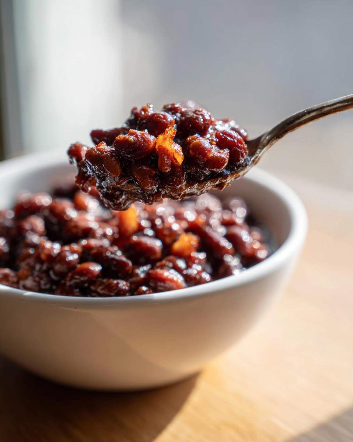 A spoonful of rich, dark, homemade Baked Beans lifted above a white bowl.
