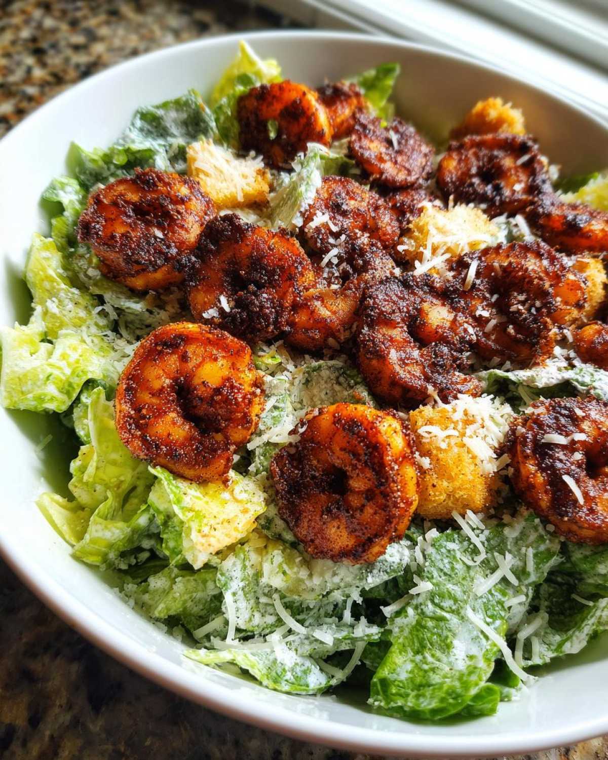 A close-up view of a Blackened Shrimp Caesar Salad topped with blackened shrimp, croutons, and grated Parmesan cheese.