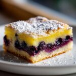 A close-up of a single square slice of Blueberry Lemon Pie Bars topped with powdered sugar on a white plate.