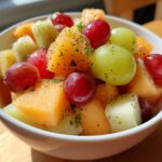 Close-up of a vibrant bowl of Fresh Fruit Salad featuring cantaloupe, kiwi, and red and green grapes sprinkled with poppy seeds.