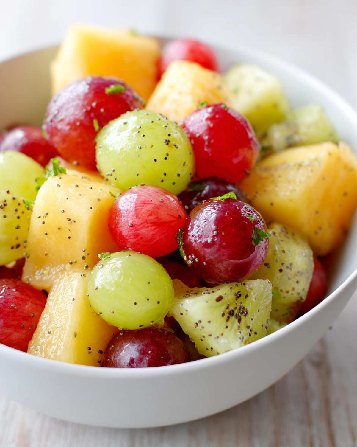 Close-up of a white bowl filled with vibrant Boat Snacks: Fresh Fruit Salad including red and green grapes, cantaloupe, and kiwi, sprinkled with poppy seeds.
