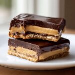 Two stacked squares of rich buckeye graham crackers bars featuring a graham cracker crust, peanut butter filling, and chocolate topping.