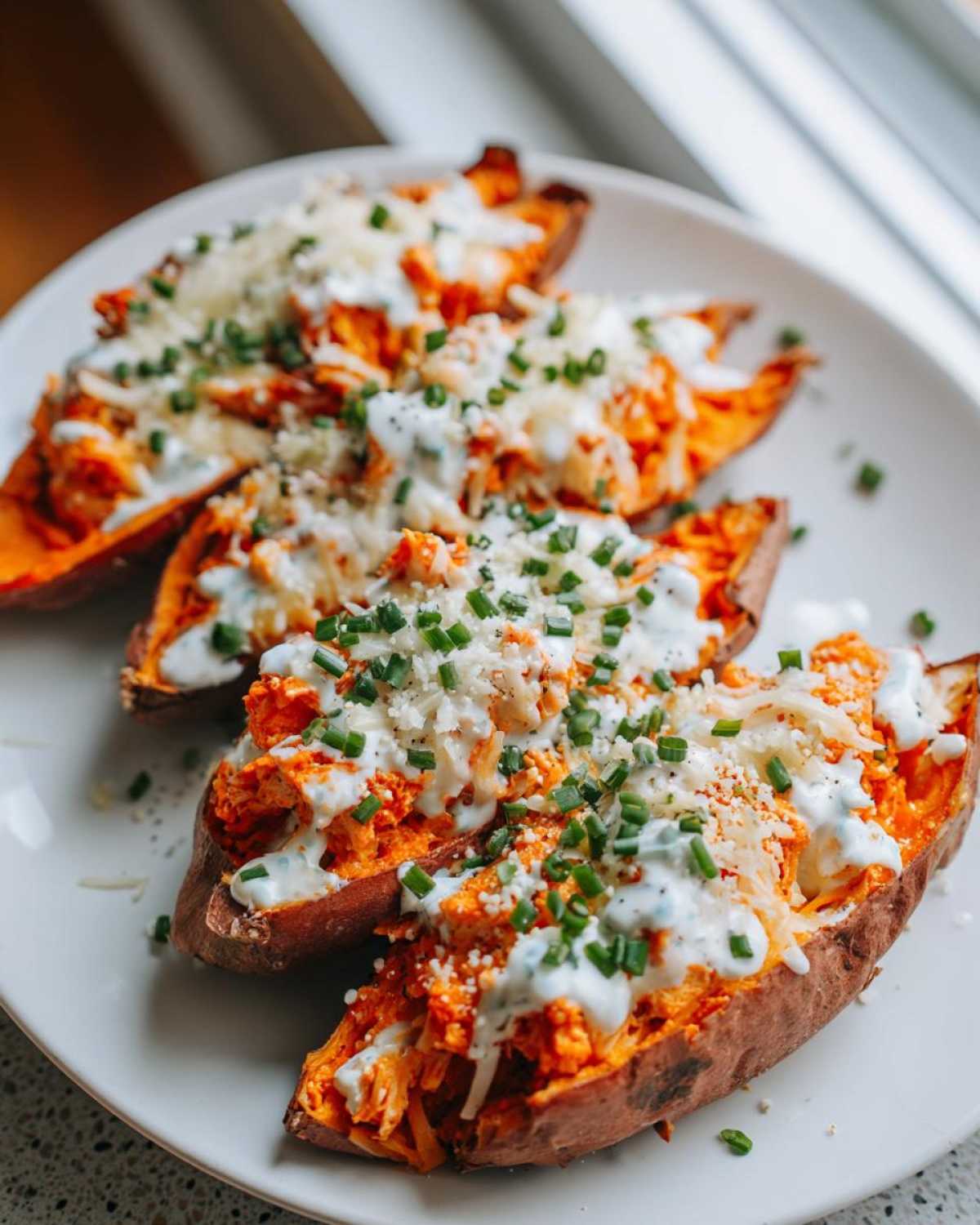Four halves of Buffalo Chicken Stuffed Sweet Potatoes topped with melted cheese, ranch dressing, and chives on a white plate.