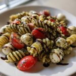 A close-up of a serving of caprese pasta salad featuring rotini pasta, cherry tomatoes, mozzarella balls, and pesto, drizzled with balsamic glaze.