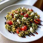 A close-up of a serving of caprese pasta salad featuring fusilli pasta, cherry tomatoes, mozzarella balls, and pesto, drizzled with balsamic glaze.