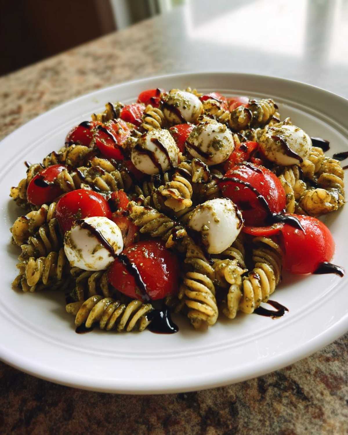 A vibrant plate of caprese pasta salad featuring fusilli pasta coated in pesto, cherry tomatoes, small mozzarella balls, and a balsamic glaze drizzle.