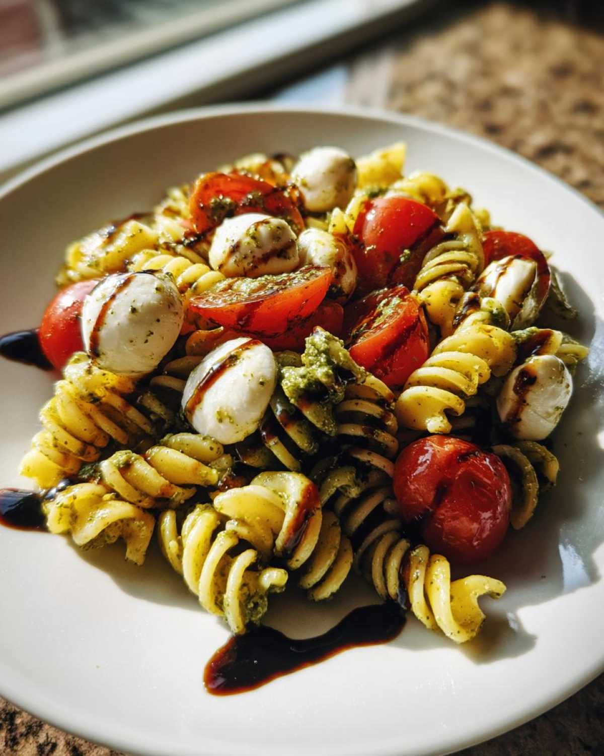 A close-up shot of a serving of caprese pasta salad featuring fusilli pasta, cherry tomatoes, mozzarella balls, and pesto, drizzled with balsamic glaze.