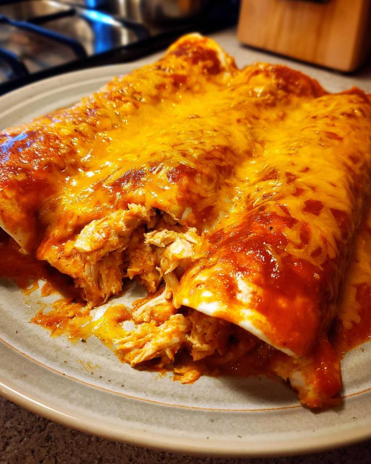 Two cheesy chicken enchiladas covered in red sauce and melted cheddar, with one showing shredded chicken filling.