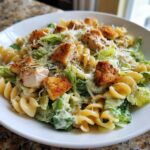 A close-up of a bowl of Chicken Caesar Pasta Salad featuring rotini pasta, romaine lettuce, grilled chicken pieces, and croutons.