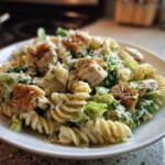 A close-up of a white plate filled with Chicken Caesar Pasta Salad featuring fusilli pasta, chicken chunks, romaine lettuce, and croutons.