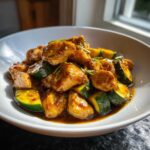 A close-up of delicious chicken zucchini stir fry coated in a savory brown sauce, served in a white bowl.