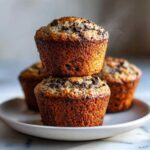 Close-up of two freshly baked chocolate chip protein muffins stacked on a white plate, with more muffins blurred in the background.