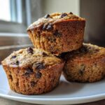 A stack of three freshly baked, warm chocolate chip protein muffins on a white plate.