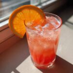 A tall glass filled with a vibrant pink Citrus Rhubarb Mocktail (Apple Juice) over ice, garnished with a bright orange slice.