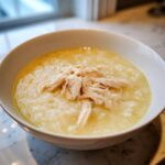 A close-up of a white bowl filled with Classic Greek Lemon Chicken Soup (Avgolemono) topped with shredded chicken.