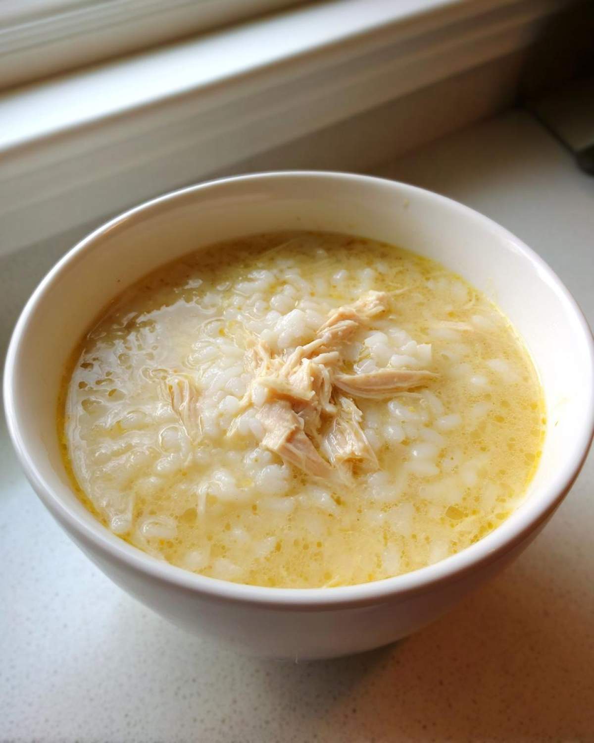 A white bowl filled with Classic Greek Lemon Chicken Soup (Avgolemono) featuring rice and shredded chicken.