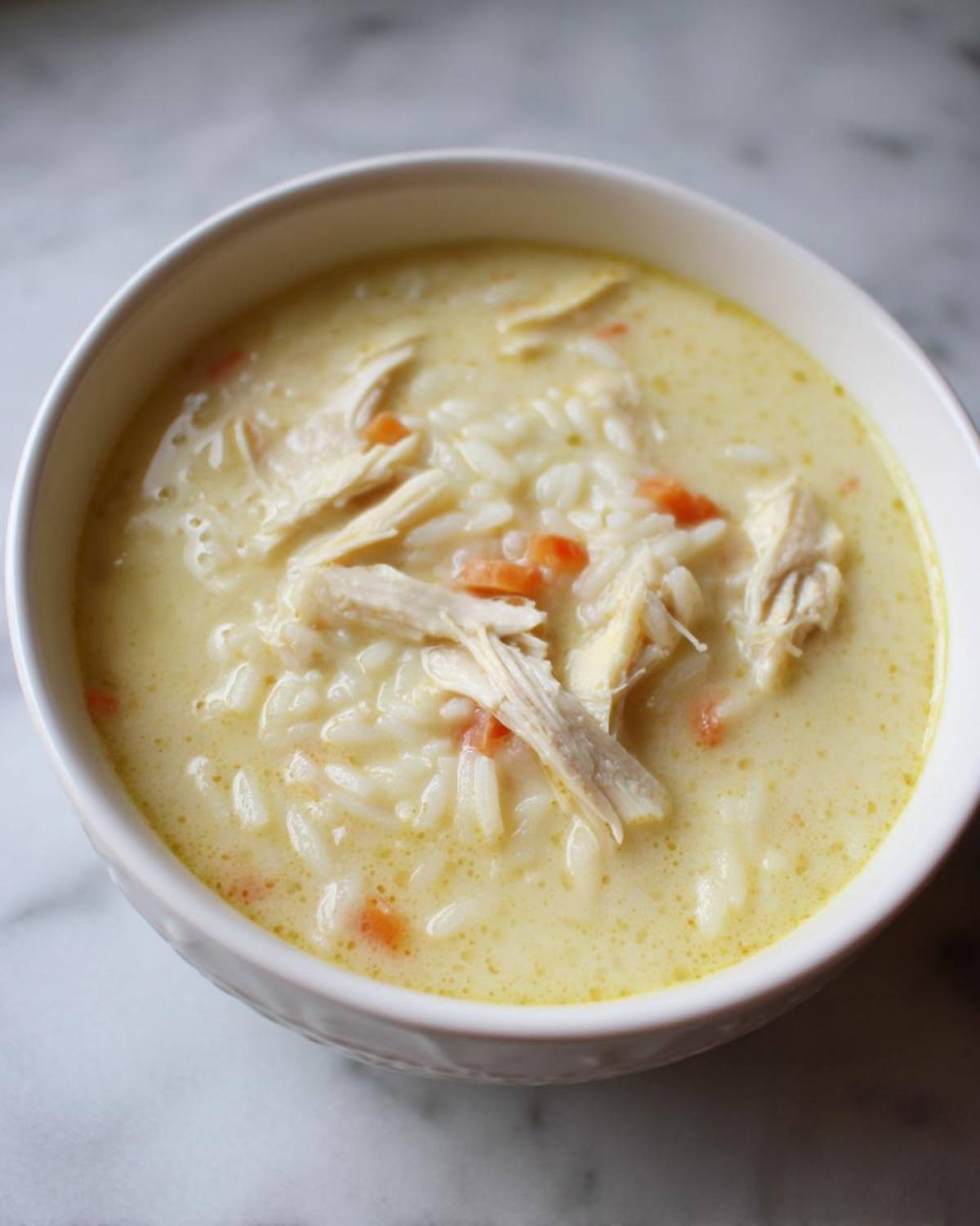 Close-up of a white bowl filled with Classic Greek Lemon Chicken Soup, showing shredded chicken, rice, and carrots in a creamy yellow broth.