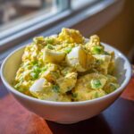 A white bowl filled with creamy Classic Mustard Potato Salad mixed with chunks of hard-boiled egg and green onions.
