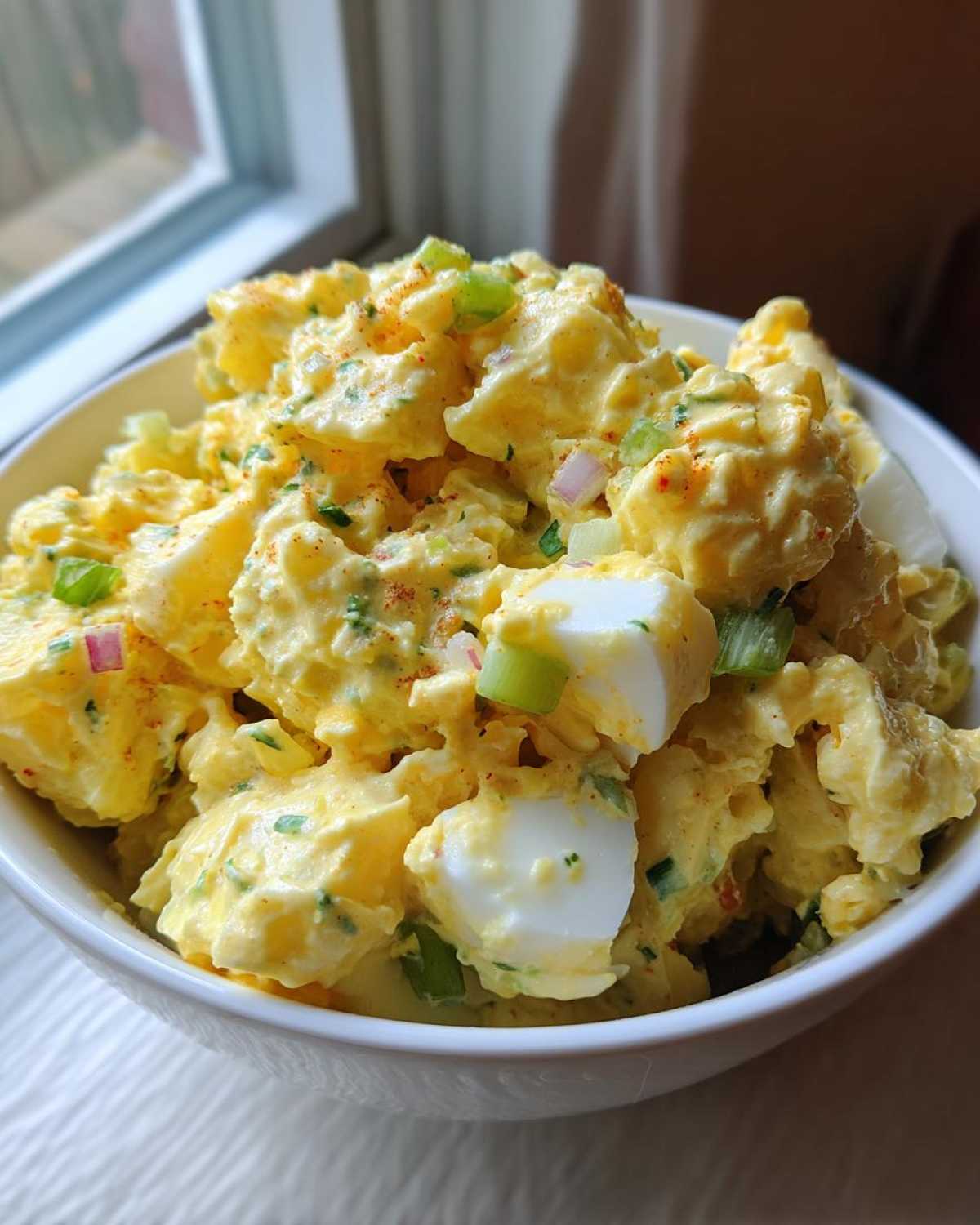 Close-up of a white bowl filled with creamy Classic Mustard Potato Salad, featuring chunks of potato, hard-boiled egg, and green onions.