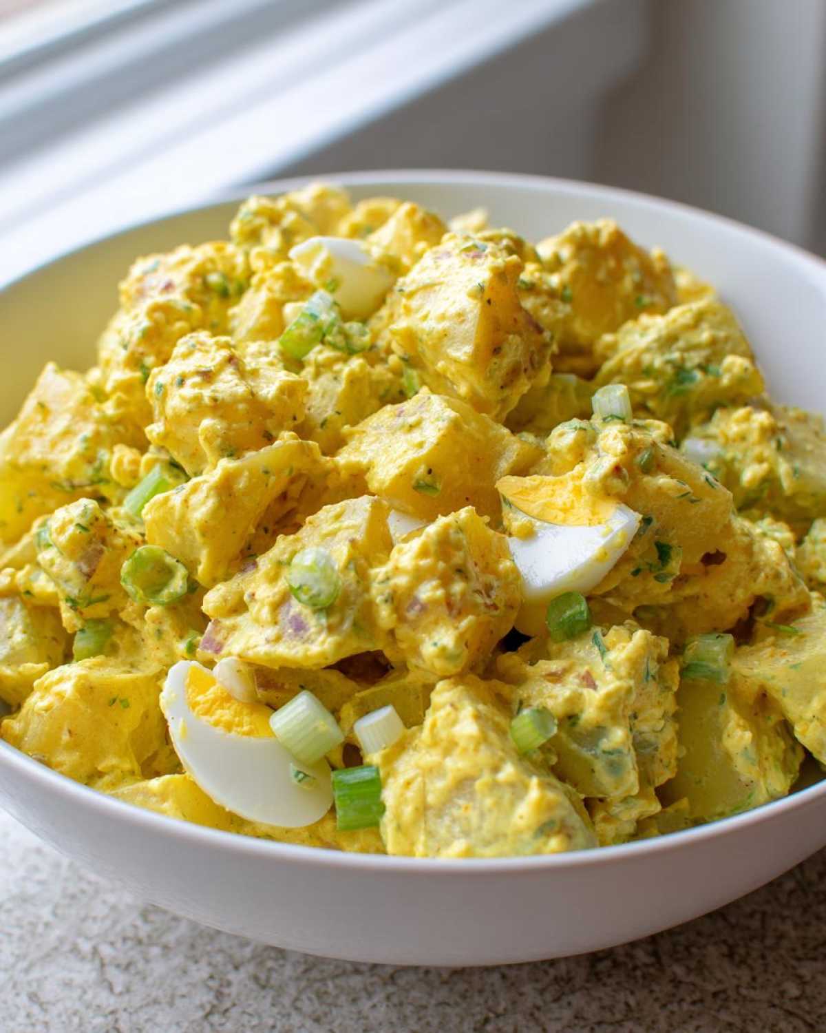 A close-up of Classic Mustard Potato Salad coated in a creamy yellow dressing, mixed with chopped hard-boiled eggs and green onions.