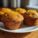 Four golden brown Classic pumpkin muffins displayed closely on a white plate, backlit by natural light.