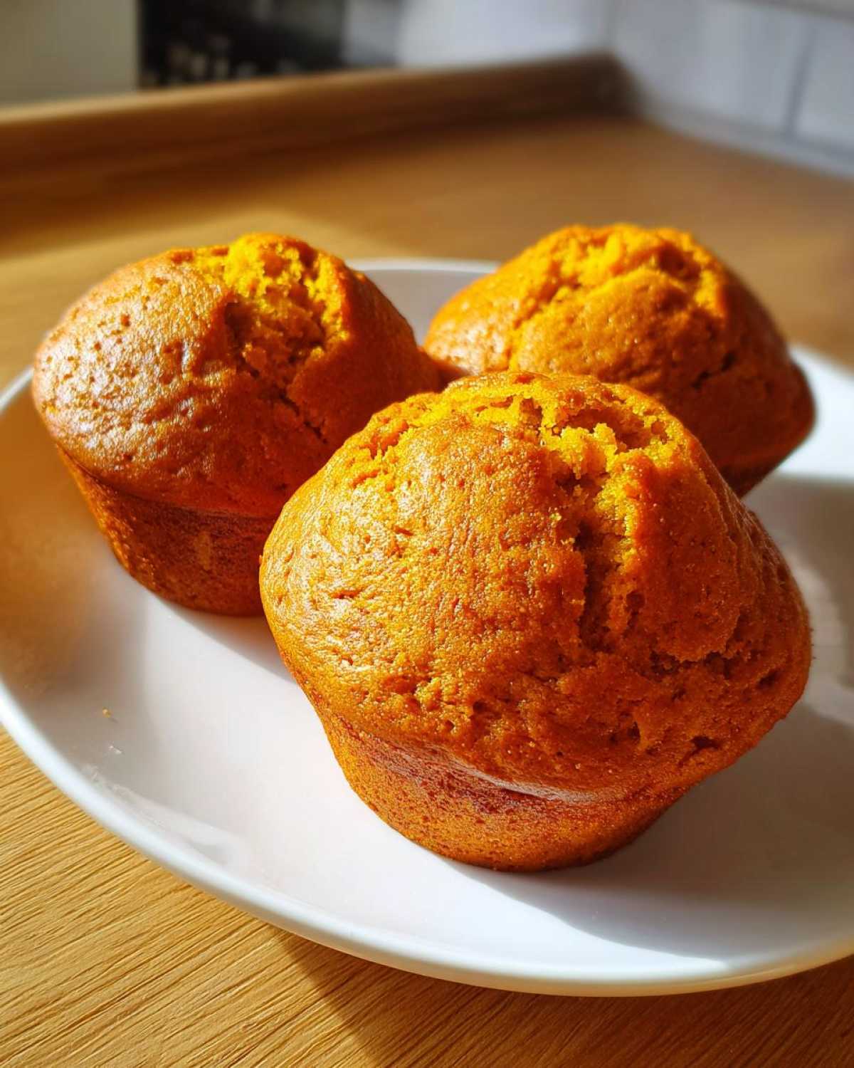 Three freshly baked Classic pumpkin muffins with a golden-orange hue sitting on a white oval plate.