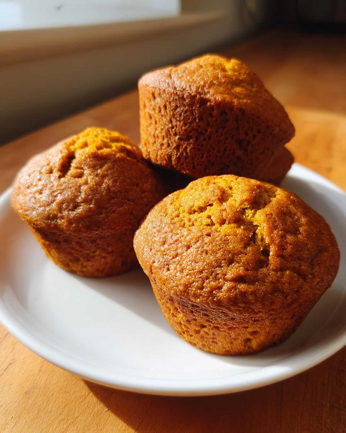 Three freshly baked Classic pumpkin muffins piled on a white plate, illuminated by sunlight.