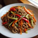 A close-up of a serving of Cold Soba Noodle Salad with Ginger Soy, tossed with julienned carrots, cucumber, and red pepper, topped with sesame seeds.