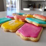 A pile of square sugar cookies topped with brightly colored, glossy sugar cookie icing in pink, yellow, blue, and orange.