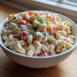 A close-up of a white bowl filled with creamy Amish macaroni salad featuring elbow pasta, red onion, celery, and peppers.