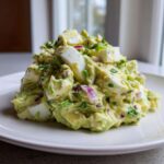 A mound of bright green Avocado Egg Salad featuring chunks of hard-boiled egg and red onion on a white plate.