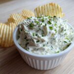 A white bowl filled with creamy Boat Dip topped with fresh chives, served with ruffled potato chips.