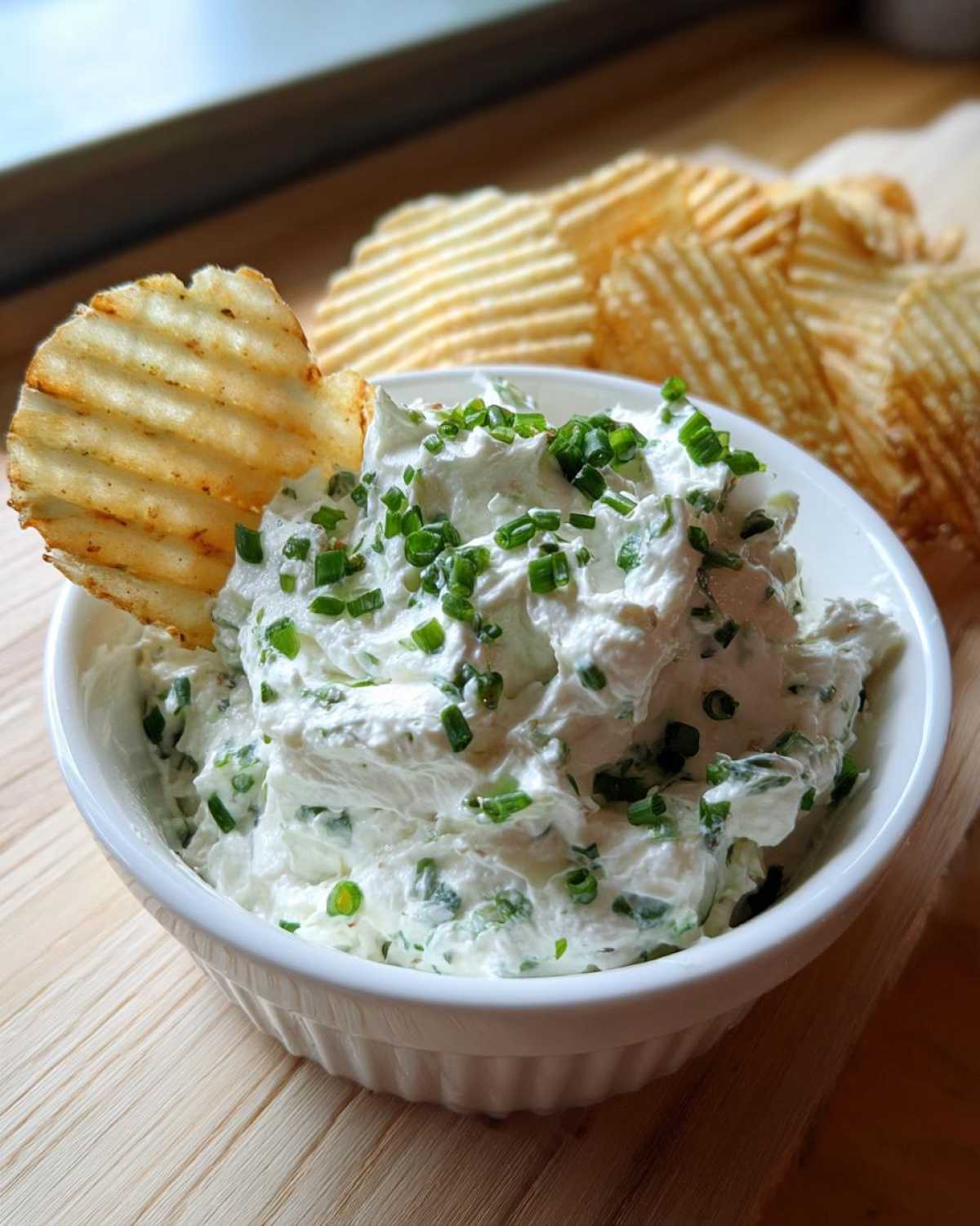 A white bowl filled with creamy Boat Dip topped with fresh chives, served with a ruffled potato chip dipped in.