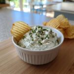 A white bowl filled with creamy Boat Dip topped with fresh chives, with a ruffled potato chip dipped in.