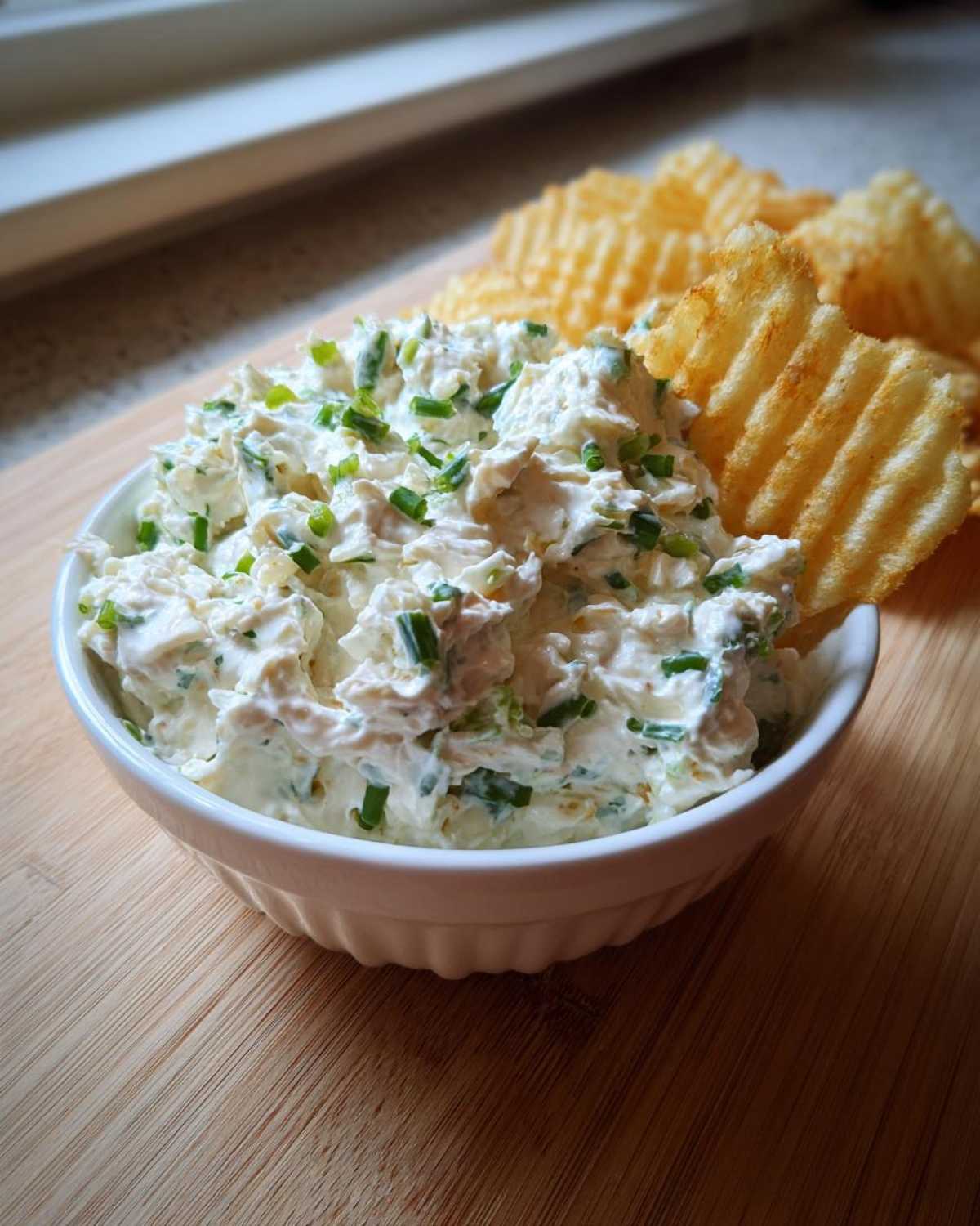 A white bowl filled with creamy Boat Dip topped with fresh chives, served next to waffle-cut potato chips.