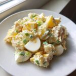 A close-up of a creamy serving of Classic Potato Salad mixed with chunks of hard-boiled egg and fresh green onions on a white plate.